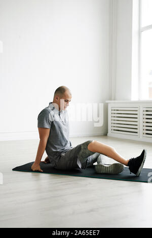 Young man with prosthesis on the leg sitting on the floor on exercise mat and exercising in the room Stock Photo