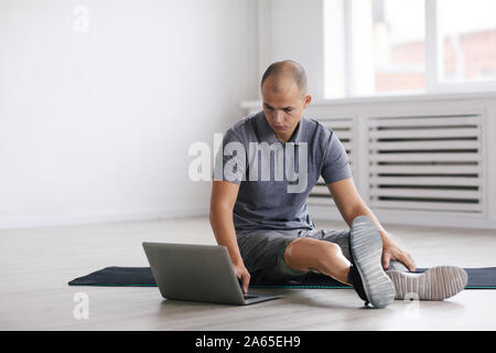 Young man with prosthesis on the leg sitting on the floor on exercise mat and using laptop computer during sports training Stock Photo