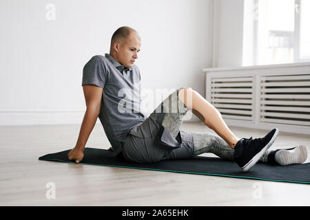 Young disabled man sitting on exercise mat and training his leg during sports training Stock Photo