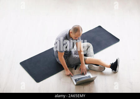 Young disabled man sitting on exercise mat and communicating online using his laptop computer in the room Stock Photo