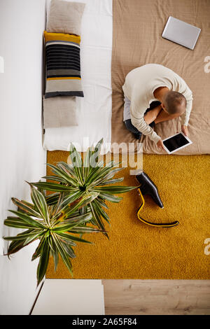 High angle view of young disabled man sitting on the bed and using digital tablet in the living room Stock Photo