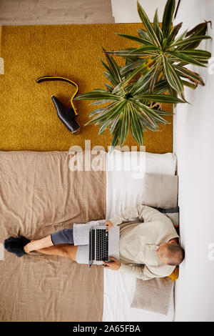 High angle view of young disabled man sitting on the bed resting and using laptop computer in his room Stock Photo