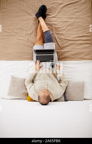 High angle view of young disabled man lying on the bed drinking coffee and working online on his laptop in the bedroom Stock Photo