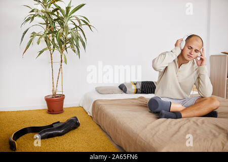 Young man with his eyes closed sitting on the bed in headphones and listening to music in his bedroom Stock Photo