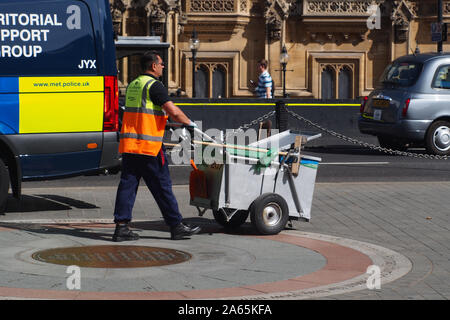 A road sweeper, street cleaner, pushing his trolley past the Houses of Parliament, Westminster,London, wearing a hi-viz jacketand two vehicles in view Stock Photo
