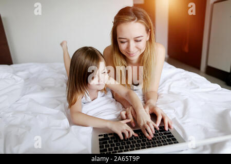 family using laptop laying on white bed woman and kid girl daughter with sunshine Stock Photo