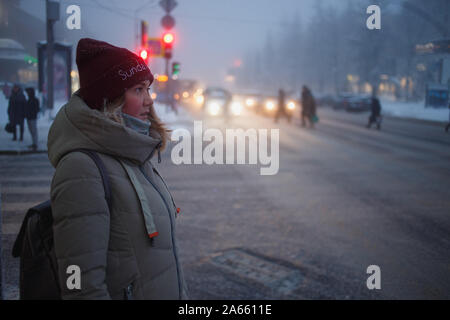 Woman walking in the city in the winter dusk time. The woman is waiting for a green traffic light signal on the crossroads. Stock Photo