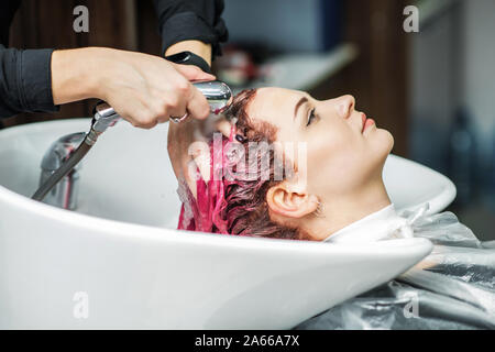 Barber is washing the girl pink hair in the beauty salon. Hands of hairdresser washes woman hair in sink at beauty salon close up. Stock Photo