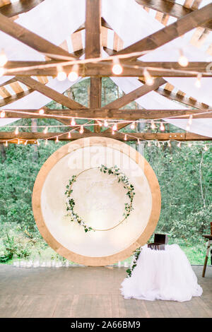 Wedding round arch in rustic style decorated with wreath from flowers and greenery, retro light bulbs on the ceiling. Beautiful wedding set up. Area Stock Photo