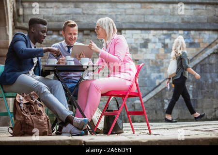 Two businessmen and a businesswoman sitting at an outdoor table in a city having a hot drink while discussing work on a digital tablet. Stock Photo