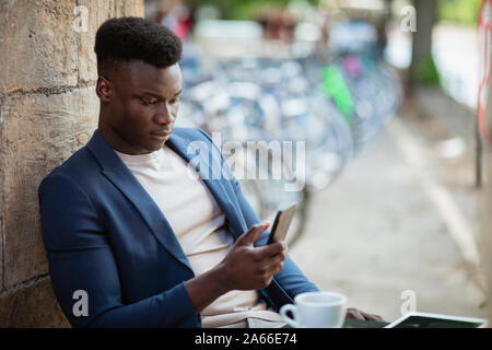 Businessman sitting down at an outdoor table in a city while typing on his mobile phone. Stock Photo