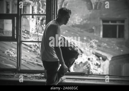 A sad guy in sports clothes and with a backpack in his hands stands on the windowsill, on which are the shards of glass, an abandoned building. Stock Photo