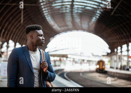 A businessman waiting for a train on the platform in a train station. He is holding a backpack over his shoulder. Stock Photo