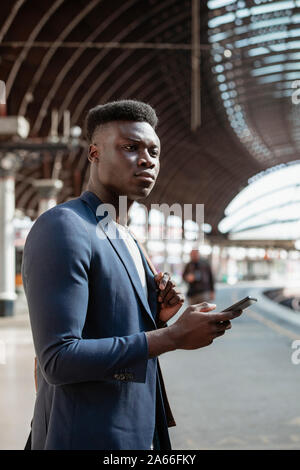 A businessman waiting for a train on the platform in a train station. He is holding a backpack over his shoulder while also holding a mobile phone in Stock Photo