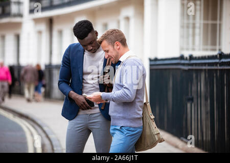 Two businessmen standing on the pavement of a city street while looking at one of their phones. Stock Photo