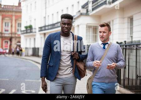 Two businessmen walking, on their way to work. They are talking and one man is holding a mobile phone ready to use it. Stock Photo