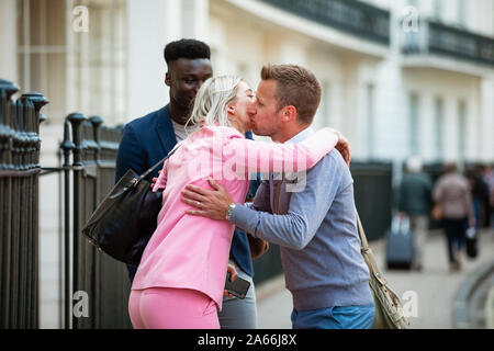 Two male and one female co-workers meeting up before work and greeting each other with a kiss on the cheek and hug. Stock Photo