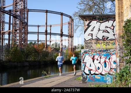 Two joggers passing empty gasometers and graffiti along the towpath of the Regent's canal near Cat and Mutton Bridge, Hackney, East London, London, UK Stock Photo