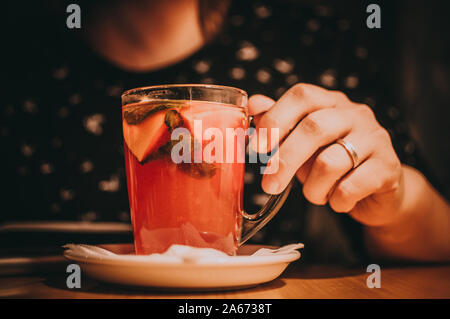 girls hand reaches for a cup with fruit tea, slices of lemon, orange and mint. A cup of tea is on a wooden table in a cafe. The concept of a cozy time Stock Photo