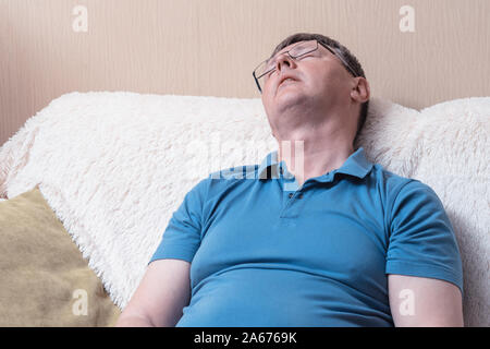 White middle-aged man fell asleep sitting on a sofa. A man with glasses and a blue T-shirt sleeps while sitting Stock Photo