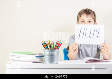 Sad tired frustrated boy sitting at the table with many books and holding paper with word Help. Learning difficulties, education concept. Stock Photo