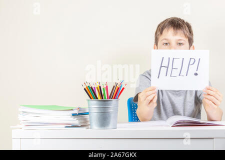 Sad tired frustrated boy sitting at the table with many books and holding paper with word Help. Learning difficulties, education concept. Stock Photo