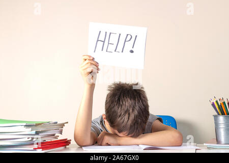 Sad tired frustrated boy sitting at the table with many books and holding paper with word Help. Learning difficulties, education concept. Stock Photo