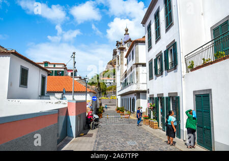 Ribeira Brava, Madeira, Portugal - Sep 9, 2019: Street in the historical center of Madeiran city with Roman Catholic church in the background. People on the streets. Historical houses. Sunny day. Stock Photo