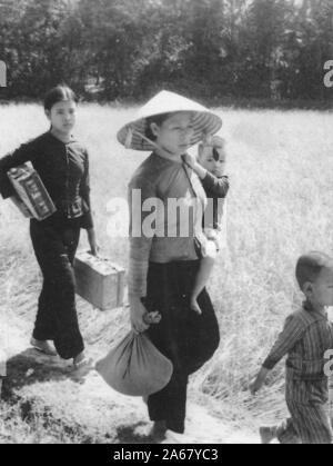 Angled full-length shot of a young boy, a woman holding a toddler and a bag, and a teenage girl carrying suitcases, walking on a dirt path on a sunny day, Vietnam, 1965. () Stock Photo