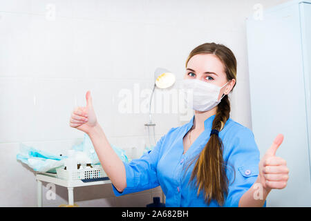 Attractive female doctor in mask woman show thumbs up hand sign with hospital background Stock Photo