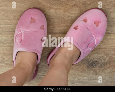 Women feet are shod in a home pink slippers. Cozy, warm and comfortable domestic shoes. On a brown wooden floor. Top view. Stock Photo