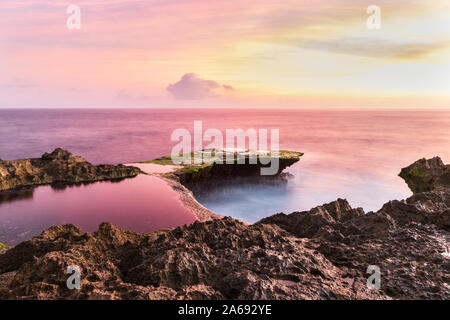 Devil's Tear at sunset, island of Nusa Lembongan, Bali, Indonesia. Rocky shore in foreground. Tidepool and ocean pink from setting sun's reflection. Stock Photo