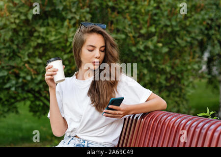 Beautiful teenage girl drinks coffee in a paper cup and flips through news feed in her smartphone. Coffee to go. Takeaway coffee. Vacation, travel. Su Stock Photo
