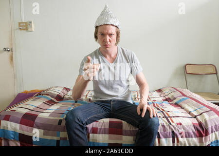 Angry young man with tin foil hat pointing at camera in the bedroom Stock Photo
