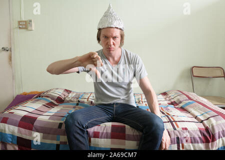 Stressed young man with tin foil hat giving thumbs down in the bedroom Stock Photo