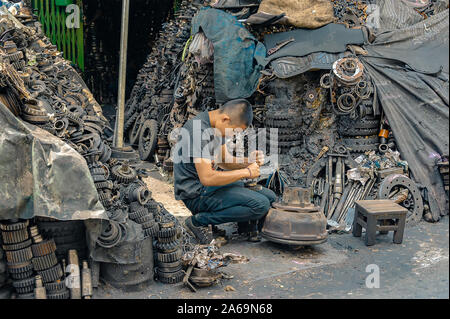 Bangkok - September 28, 2019: A worker dismantles an engine into pieces to sell as spare parts with mountain of engine pieces surrounding him. Stock Photo