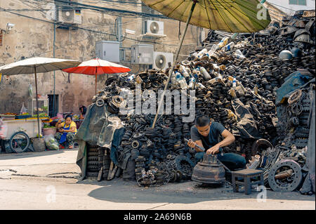 Bangkok - September 28, 2019: A worker dismantles an engine into pieces to sell as spare parts with mountain of engine pieces surrounding him. Stock Photo