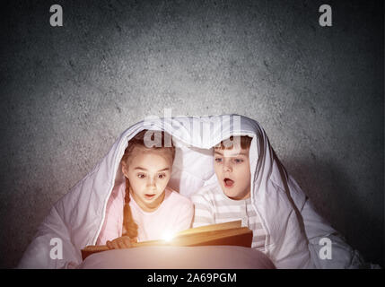 Surprised children reading scary stories in bed before going to sleep. Amazed girl and boy in pajamas hiding under blanket together. Covered kids with Stock Photo