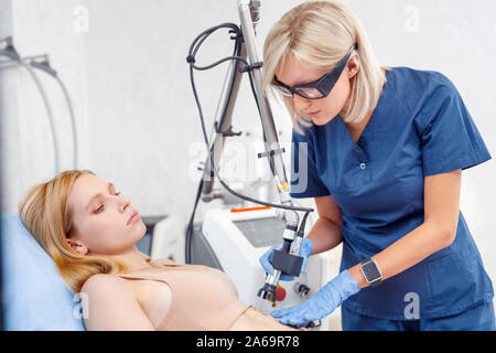 Cosmetology Service. Young woman at beauty clinic lying while doctor in safety goggles using CO2 laser removing mole concentrated Stock Photo