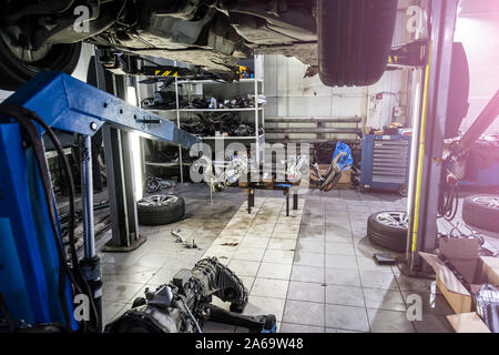 An old used car without wheel, raised on a lift for repair and under it a detached engine suspended on a blue crane near workbench in a vehicle repair Stock Photo