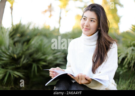 Chinese smiling girl sitting on stump in park and reading papers. Stock Photo