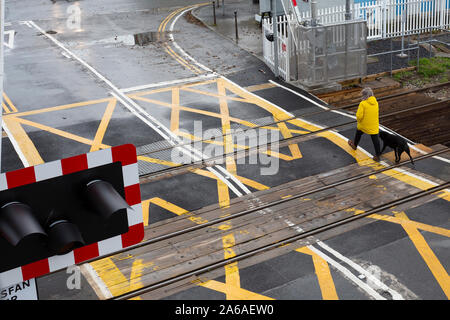 Woman in yellow coat walks across a clear railway level crossing after the barriers have been raised Stock Photo