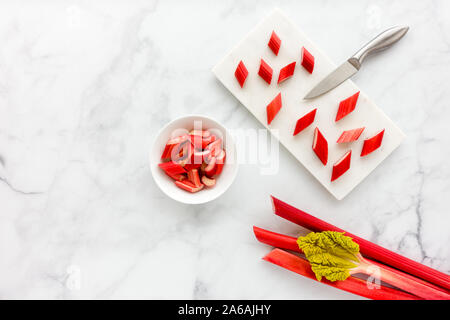 Cut young forced rhubarb pieces in a white porcelain bowl and on marble cutting board with knife on white marble background with copy space. Top view. Stock Photo