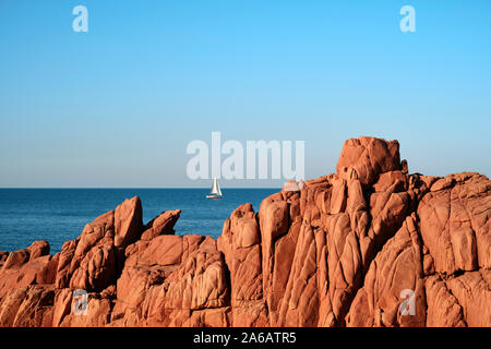 A yacht sailing and Rocce rosse / Porphyry Red Rocks Beach, Arbatax, Ogliastra coast in Tortolì, Sardinia, in Italy Stock Photo