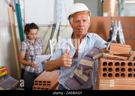 Happy experienced elderly bricklayer posing near red brick stack in building under construction Stock Photo