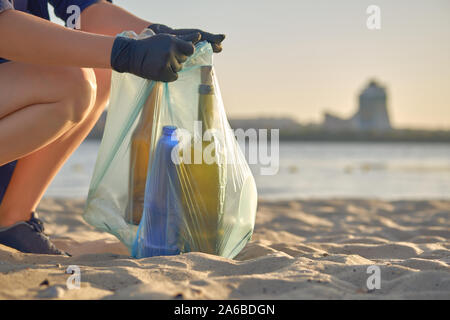 Young volunteer in black gloves is walking with garbage bag along a dirty beach of the river and cleaning up trash, used glass and plastic bottles. Pe Stock Photo