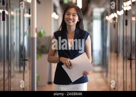 Portrait of smiling Asian businesswoman holding document, standing in hallway Stock Photo
