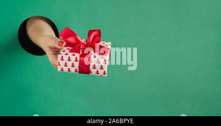 Female hand holding Christmas gift box through round hole in green paper. Cose up. Xmas holiday. Sale. Boxing day. Stock Photo
