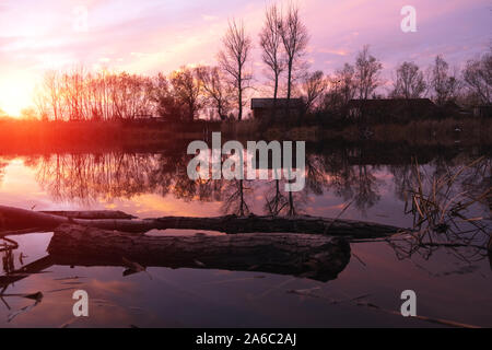 Silhouettes of houses and trees of an abandoned village on the lake against a bright scarlet, autumn sunset. Stock Photo