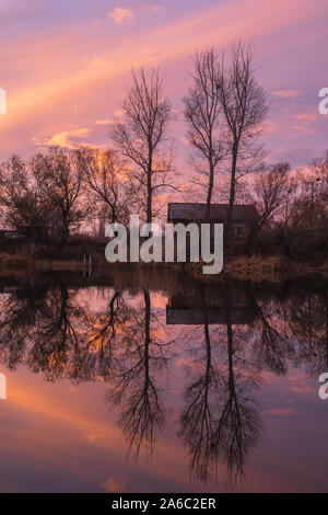 Silhouettes of houses and trees of an abandoned village on the lake against a bright scarlet, autumn sunset. Stock Photo